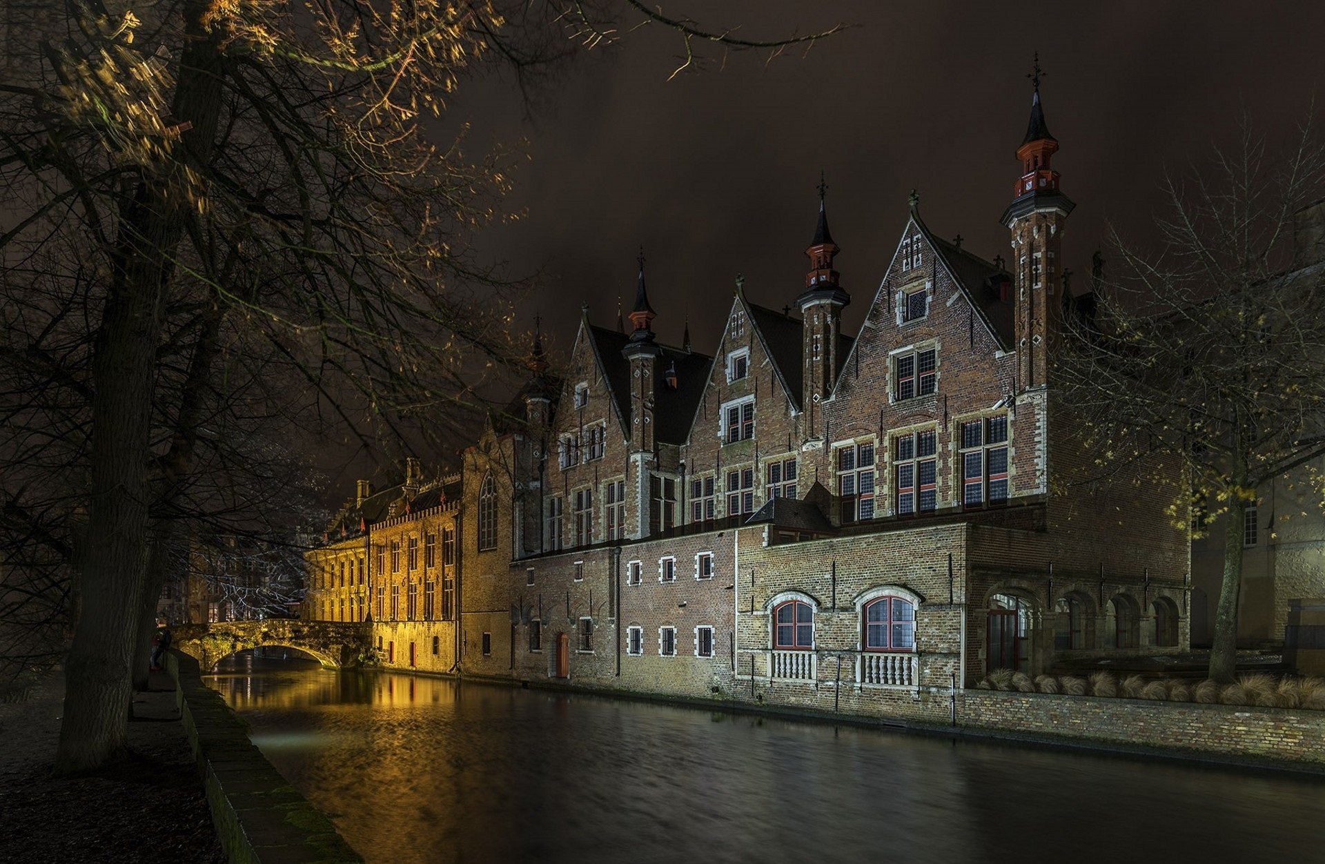 A nighttime HD desktop wallpaper of man-made historic buildings along a canal in Bruges, softly illuminated under a dark sky.