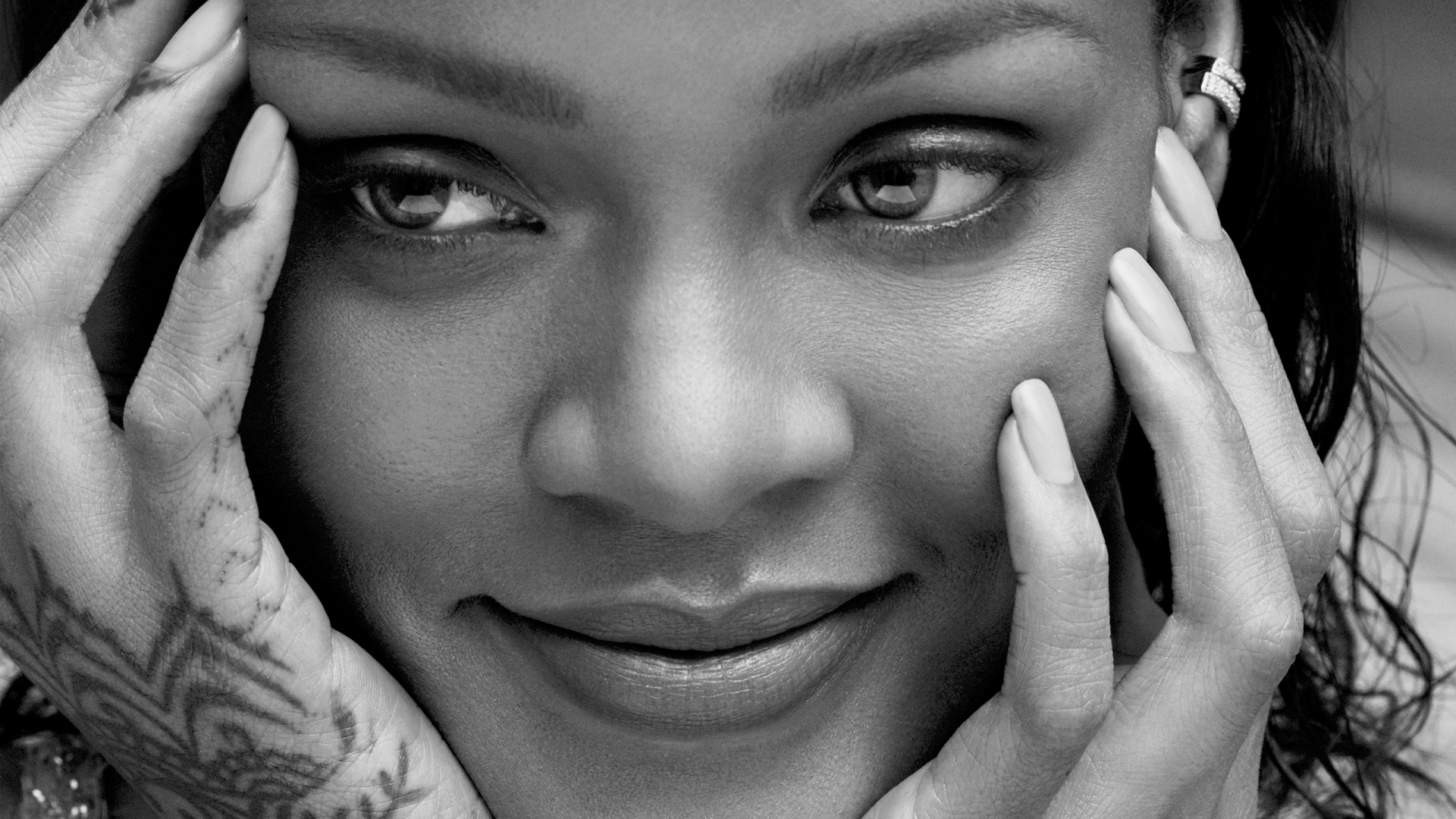 Black-and-white 2K Quad HD desktop wallpaper: close-up portrait of a smiling singer with hands framing her face, henna on one hand and an ear cuff, evoking music and style.