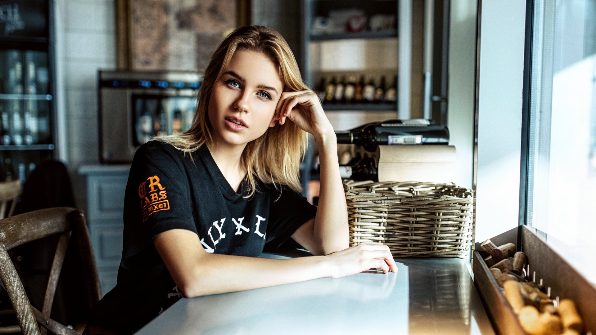 Blonde woman model with striking blue eyes sits at a bright café table, captured in sharp 4K Ultra HD quality for PC desktop wallpaper.