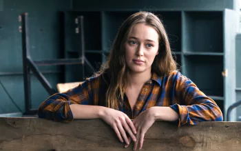 Alycia Debnam Carey, Australian actress with brunette hair, poses thoughtfully against a muted industrial background in this 4K Ultra HD desktop wallpaper.
