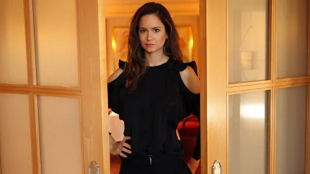 HD desktop wallpaper of a person standing between open doors, hands on hips, wearing a black outfit against a warmly lit interior background.