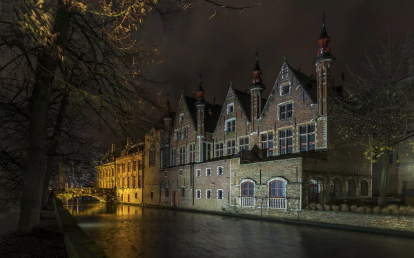 A nighttime HD desktop wallpaper of man-made historic buildings along a canal in Bruges, softly illuminated under a dark sky.