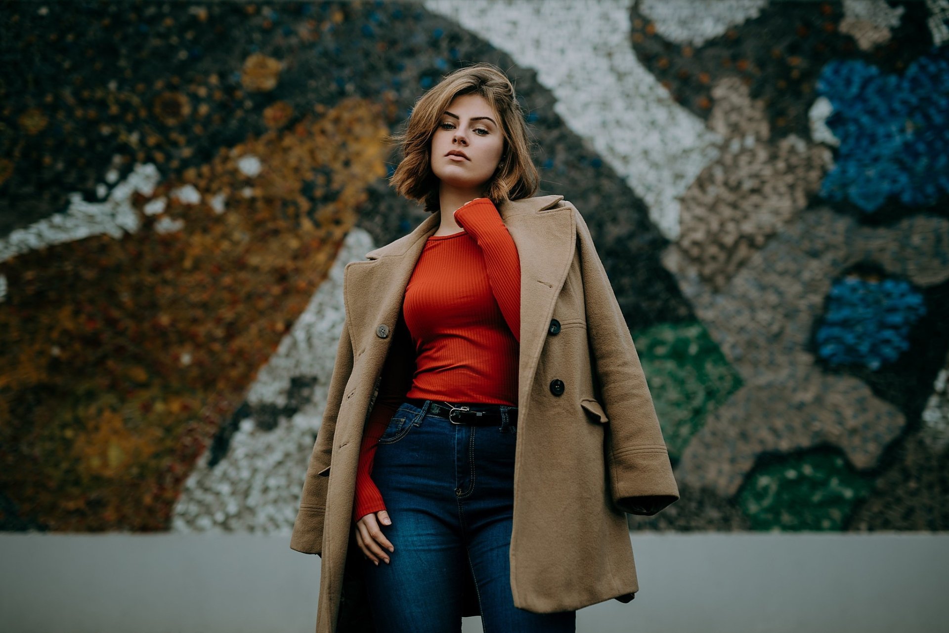 Brunette woman with short hair wearing a coat and red top stands confidently against a textured mural, captured in an HD PC desktop wallpaper image.