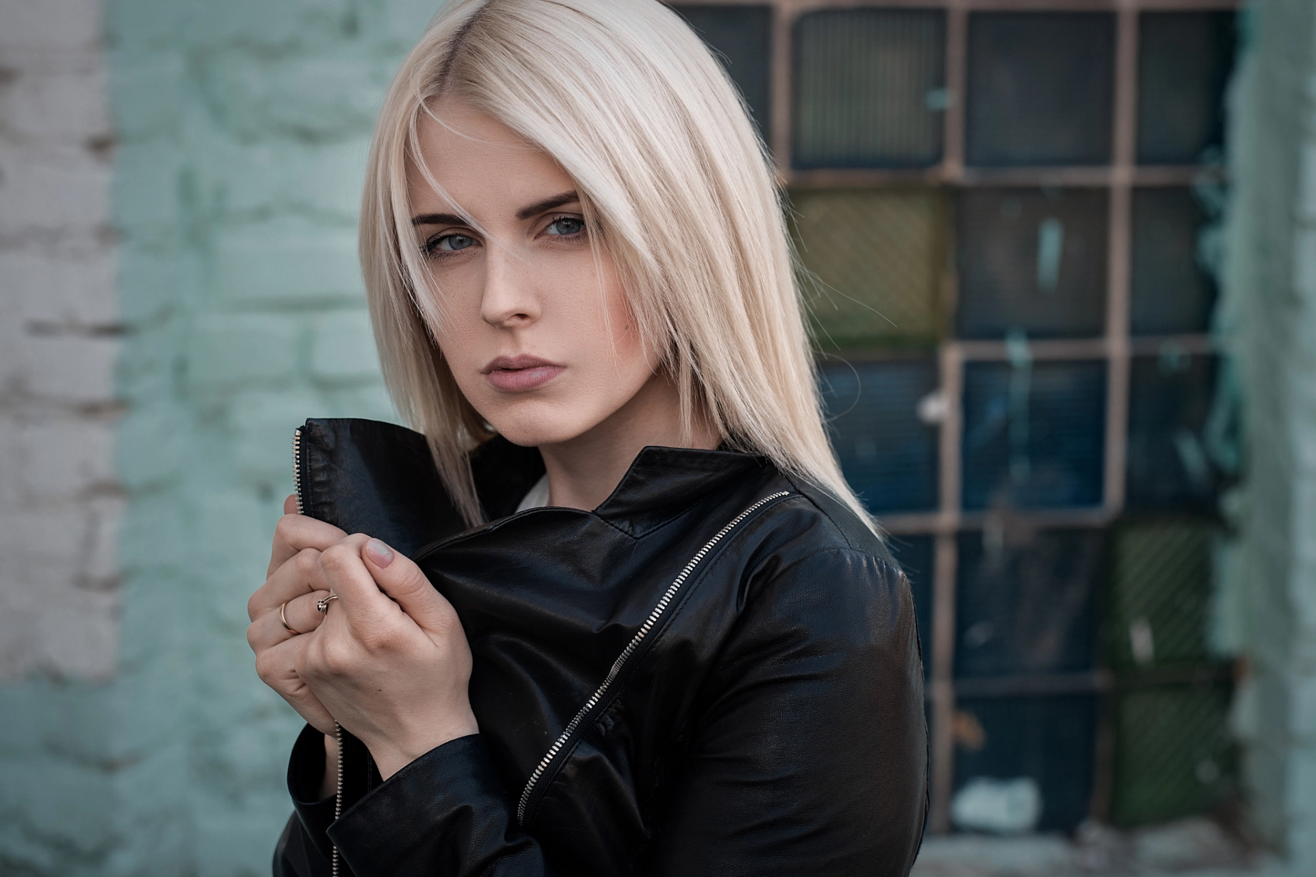 HD PC desktop wallpaper: blonde woman model with blue eyes in a black leather jacket against a weathered brick-and-window urban backdrop.