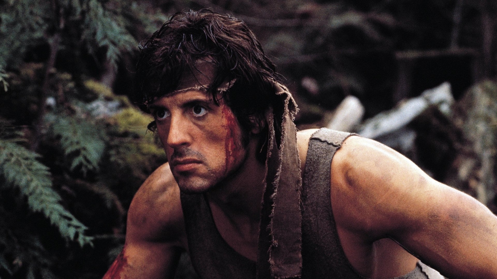 HD PC desktop wallpaper — movie still from First Blood: a bloodied, tense soldier crouches in a forest, sleeveless shirt and head bandage.