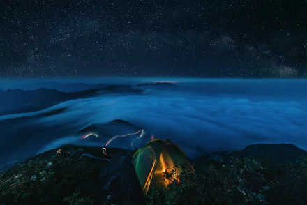 HD desktop wallpaper featuring a camping scene under a starry sky with clouds enveloping mountainous terrain, highlighting a glowing tent atop a ridge.