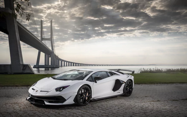 White Lamborghini Aventador SVJ supercar parked by a bridge under dramatic clouds, 5K Ultra HD desktop wallpaper of the vehicle.