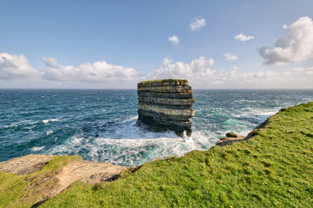 Dun Briste Sea Stack rises from the ocean off Ireland’s rugged coastline, surrounded by waves and green cliffs under a partly cloudy sky.