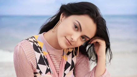 Close-up of a woman with black hair and brown eyes, wearing gold hoop earrings and a patterned sweater, resting her head on her hand by the beach — 4K Ultra HD desktop wallpaper.