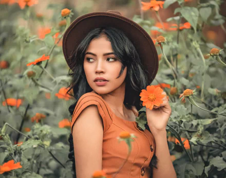 Woman model with black hair wearing a brown hat and orange dress holds an orange flower amid a flower field — 2K Quad HD PC desktop wallpaper/background.