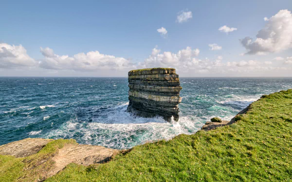 Dun Briste Sea Stack rises from the ocean off Ireland’s rugged coastline, surrounded by waves and green cliffs under a partly cloudy sky.