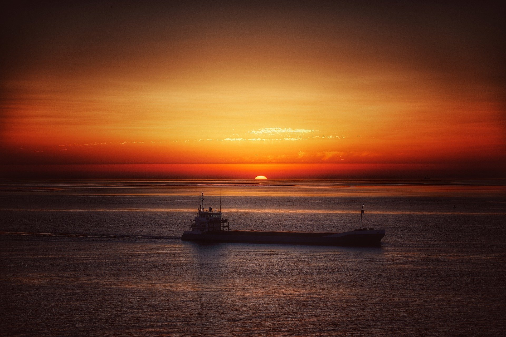 HD desktop wallpaper featuring a ship sailing on the ocean at sunset, with vibrant orange and red hues lighting up the sky and reflecting on the water’s surface.
