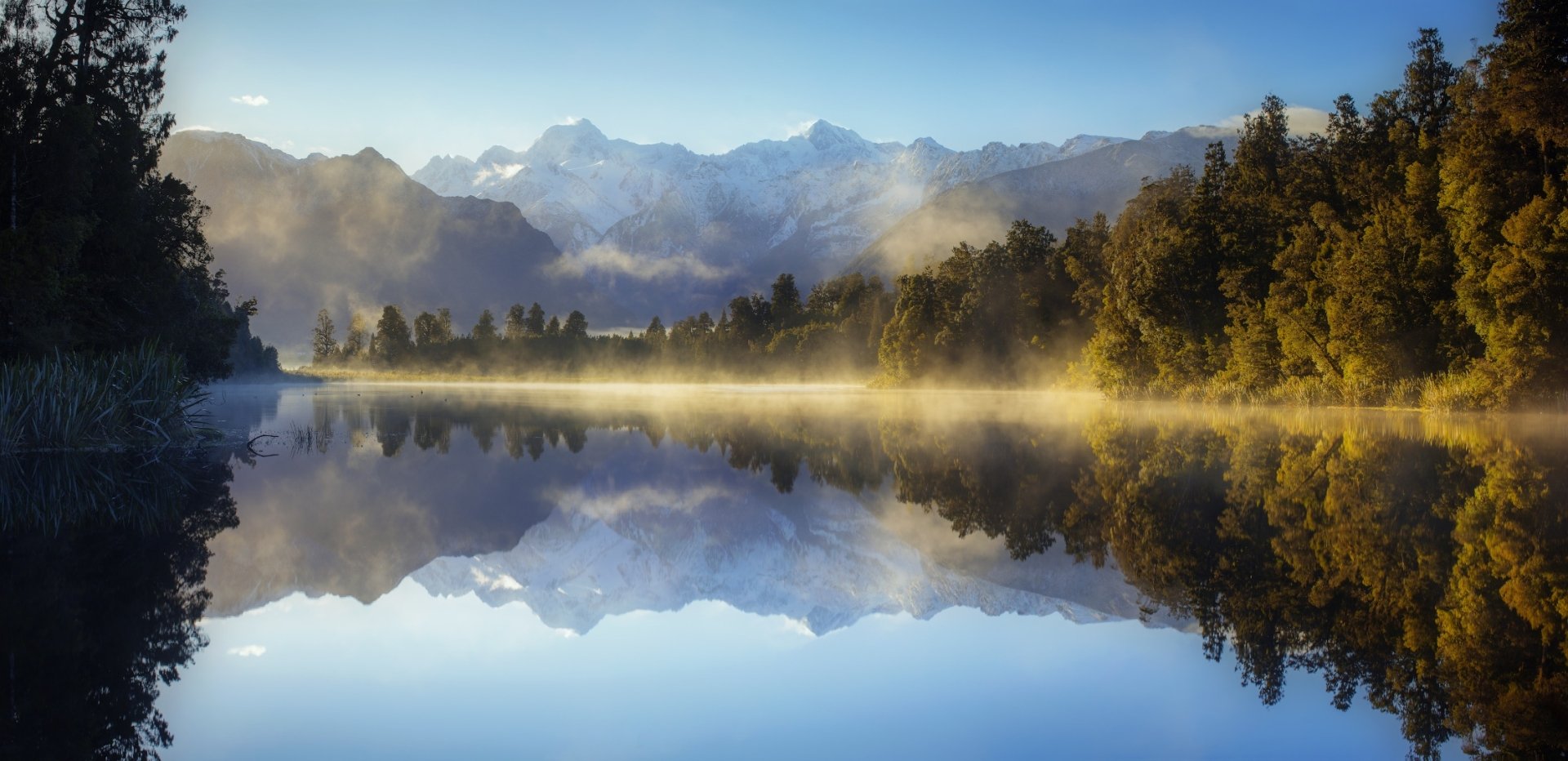 HD landscape wallpaper of a serene New Zealand lake with fog, surrounded by forest and mountains, reflecting the natural beauty in crystal-clear water.