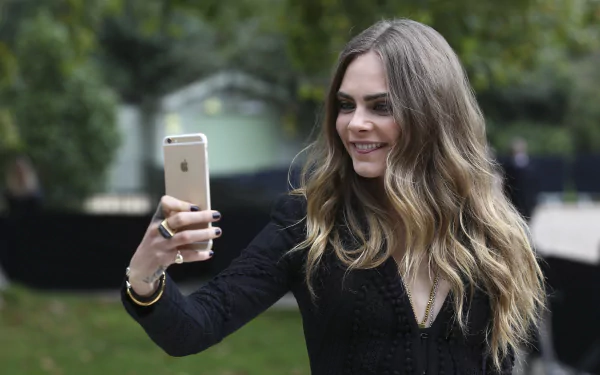 Blonde English model with long wavy hair taking a smartphone selfie, shallow depth of field, smiling portrait — 2K Quad HD PC desktop wallpaper.