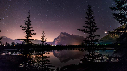HD desktop wallpaper featuring a serene lake reflecting a starlit sky and towering mountain silhouette, framed by dark trees. The starry night and tranquil nature create a breathtaking landscape.