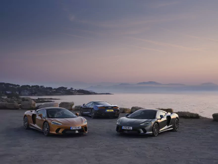 Three McLaren GT supercars — orange, silver, and gunmetal — parked on a coastal overlook at sunset, 8K Ultra HD PC desktop wallpaper.