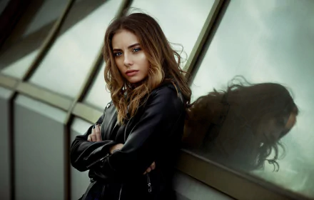 Redhead female model with blue eyes in a leather jacket leans against reflective glass — high-definition PC desktop wallpaper background.