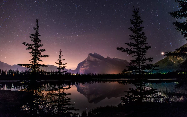 HD desktop wallpaper featuring a serene lake reflecting a starlit sky and towering mountain silhouette, framed by dark trees. The starry night and tranquil nature create a breathtaking landscape.