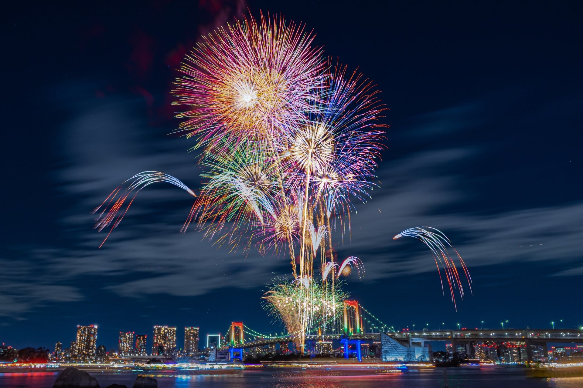 Vibrant fireworks burst over Tokyo's Rainbow Bridge at night, illuminating the city skyline in this HD desktop wallpaper capturing Japan's festive urban beauty.