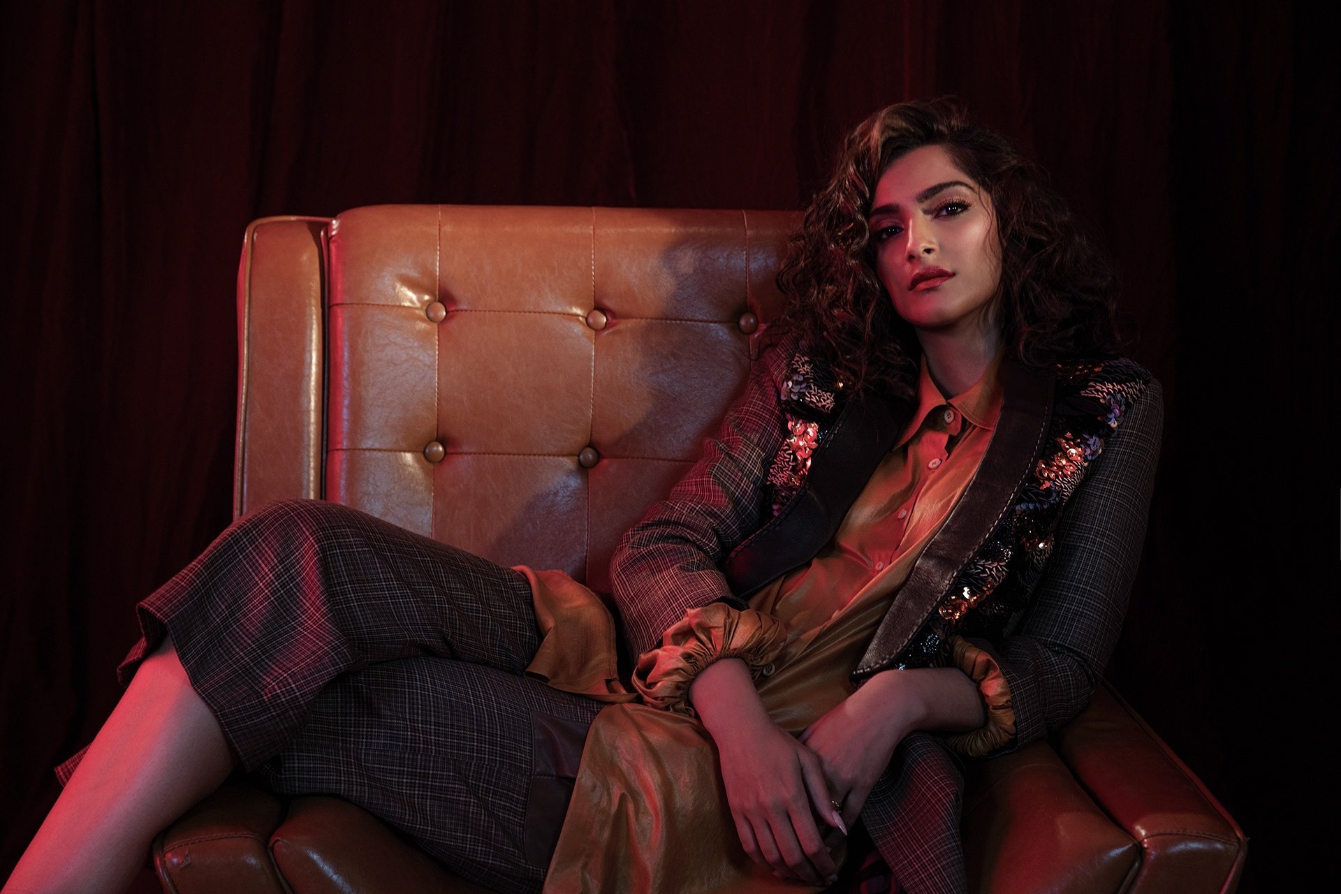 Brunette woman in a tailored coat and lipstick reclining on a tufted leather chair, moody Bollywood‑inspired lighting — HD desktop wallpaper.