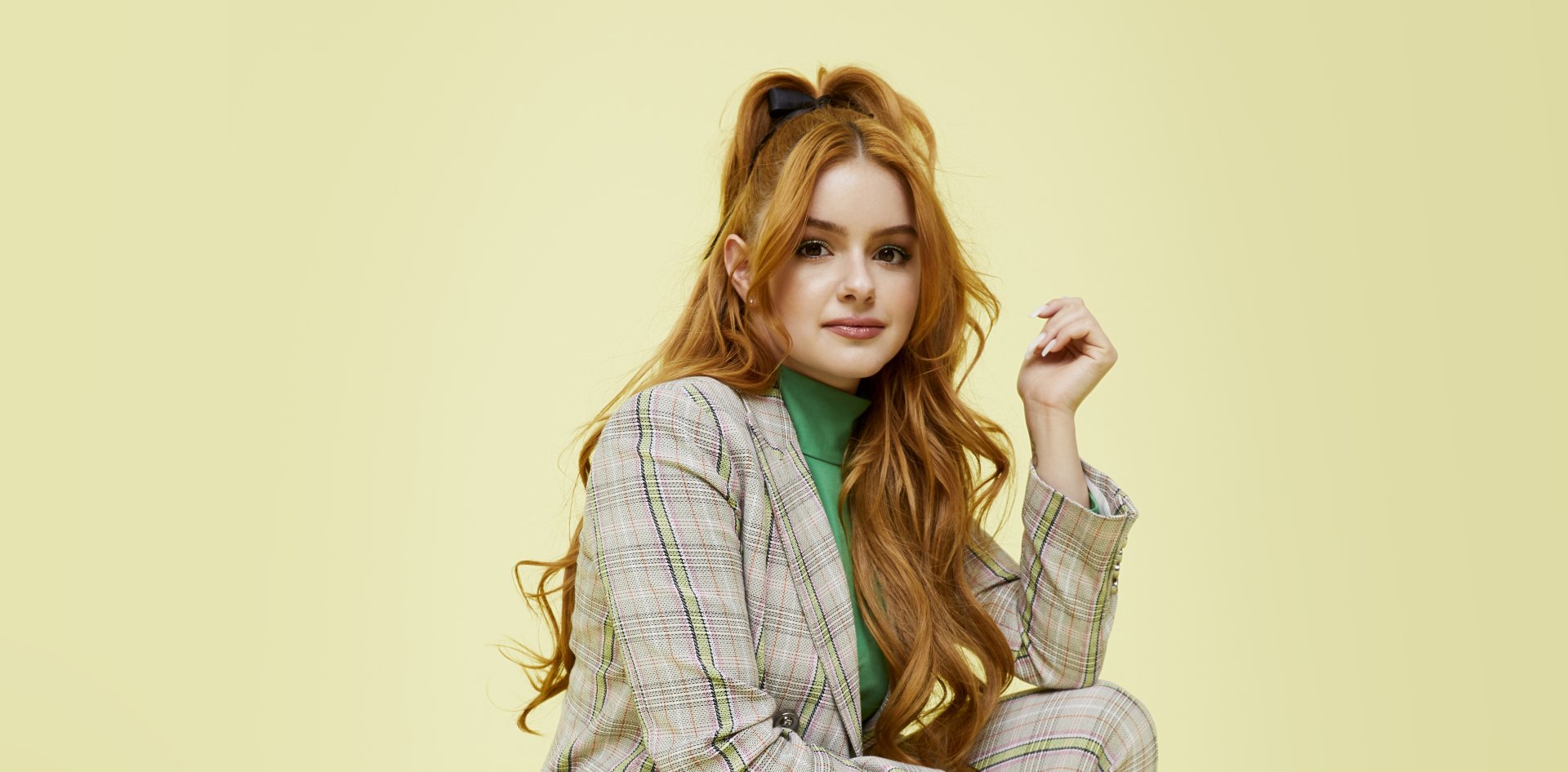 Portrait of American actress Ariel Winter with long red hair, wearing a plaid suit and green top, against a light yellow background in 4K Ultra HD quality.
