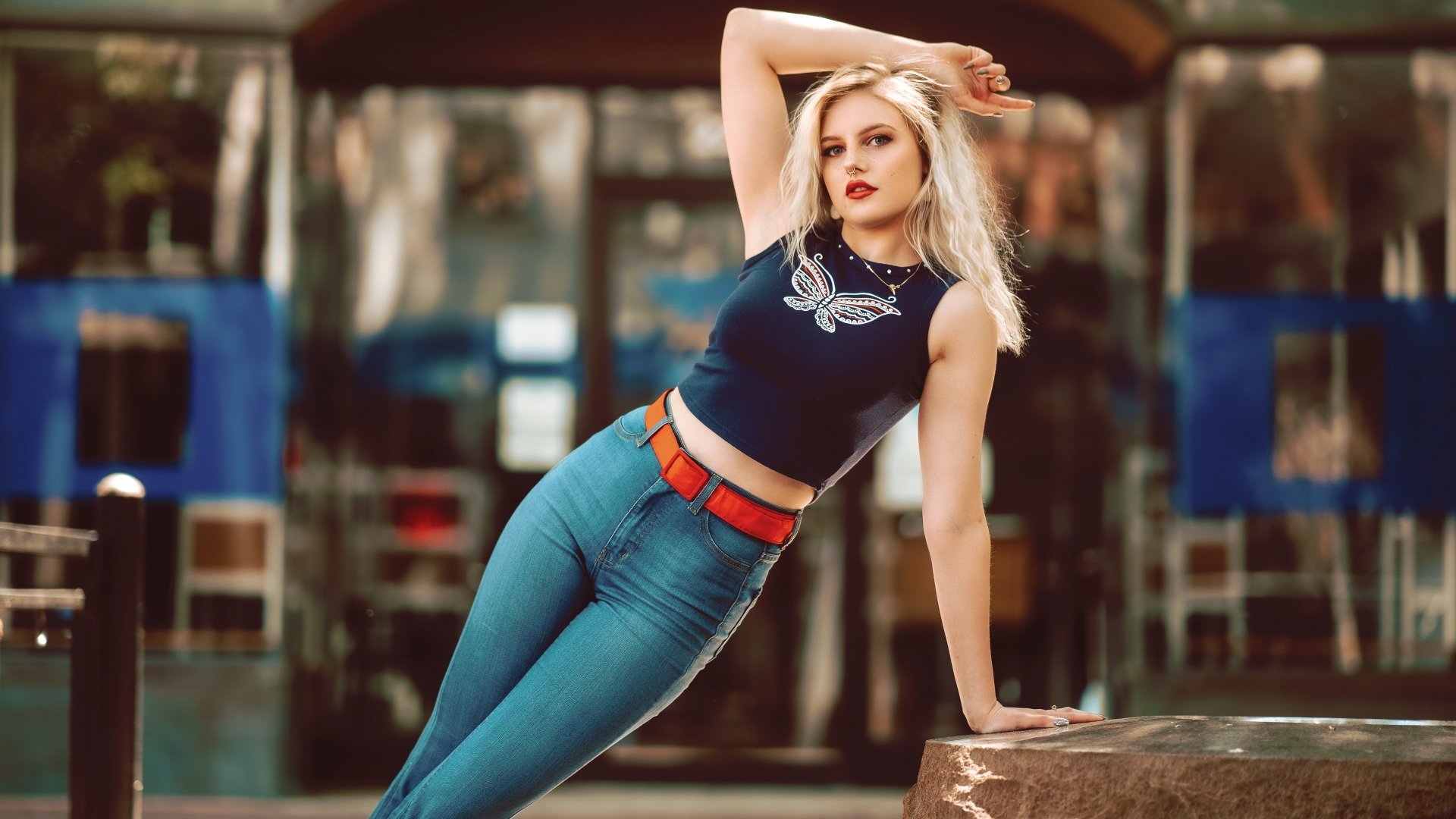 A blonde woman with blue eyes and lipstick poses in denim jeans and a sleeveless top, captured with a shallow depth of field in a 4K Ultra HD wallpaper image.