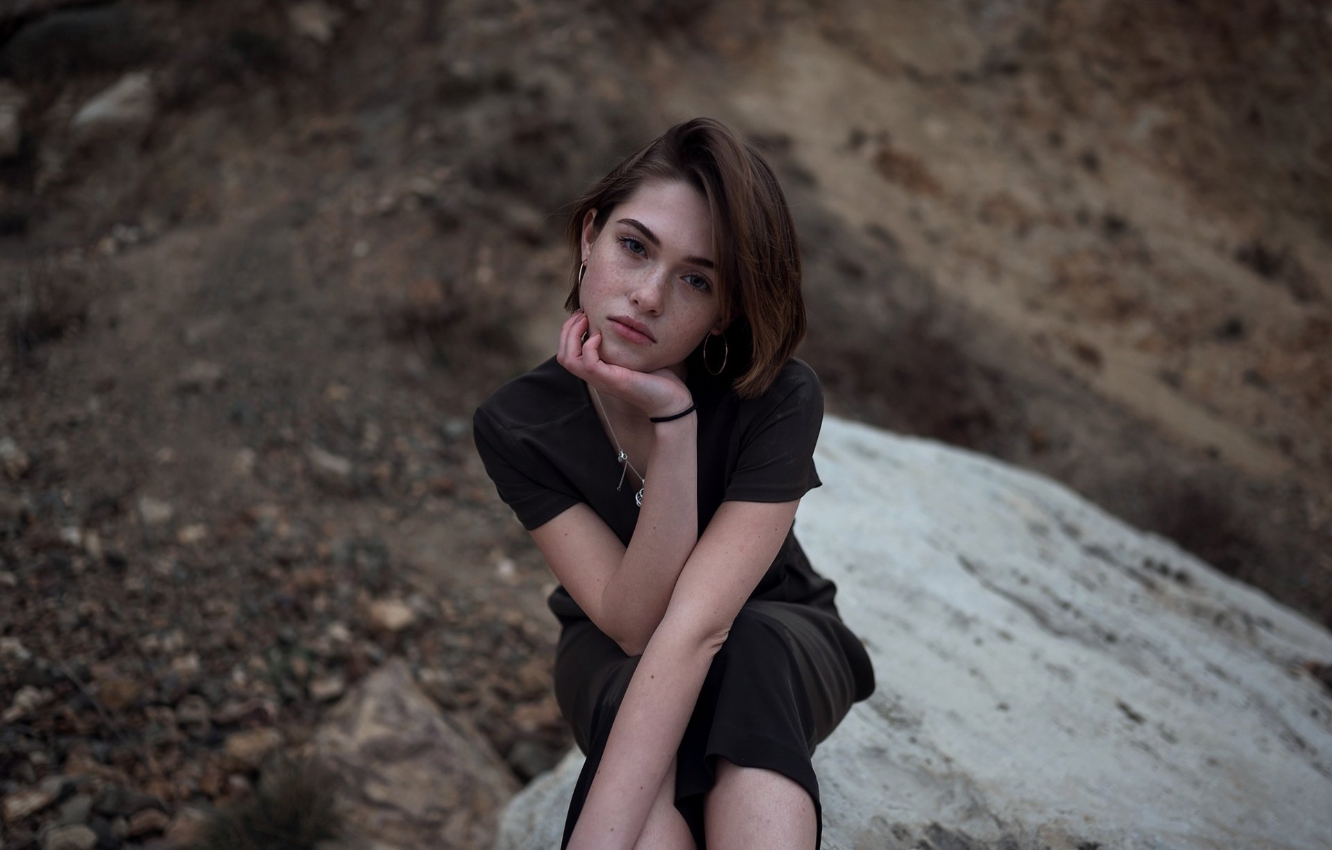 HD PC desktop wallpaper: brunette model with short hair seated on a rock, chin resting on hand, earth-toned background.