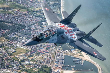 HD desktop wallpaper of a Mikoyan MiG-29 jet fighter warplane banking over a coastal town, military aircraft with clear national markings.
