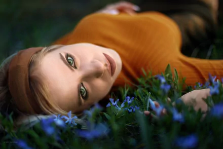 Model lying among blue flowers, staring with striking green eyes toward the camera — 2K Quad HD PC desktop wallpaper background.