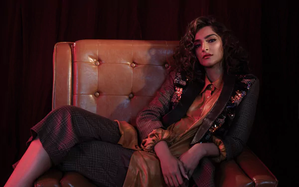 Brunette woman in a tailored coat and lipstick reclining on a tufted leather chair, moody Bollywood‑inspired lighting — HD desktop wallpaper.