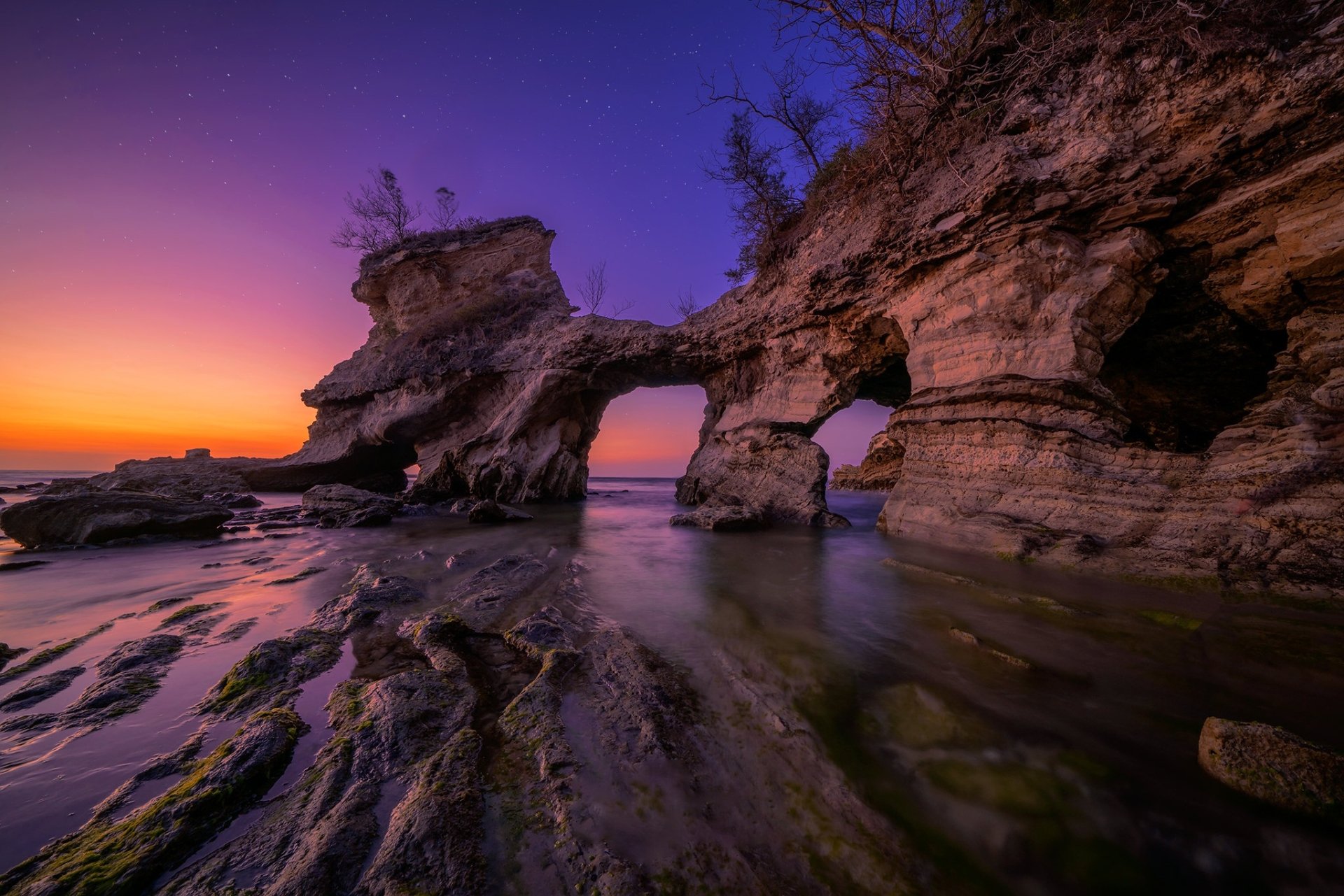 HD PC desktop wallpaper — nature coastal sunset with a stone arch, tide pools and reflective wet rock under a purple-orange sky.