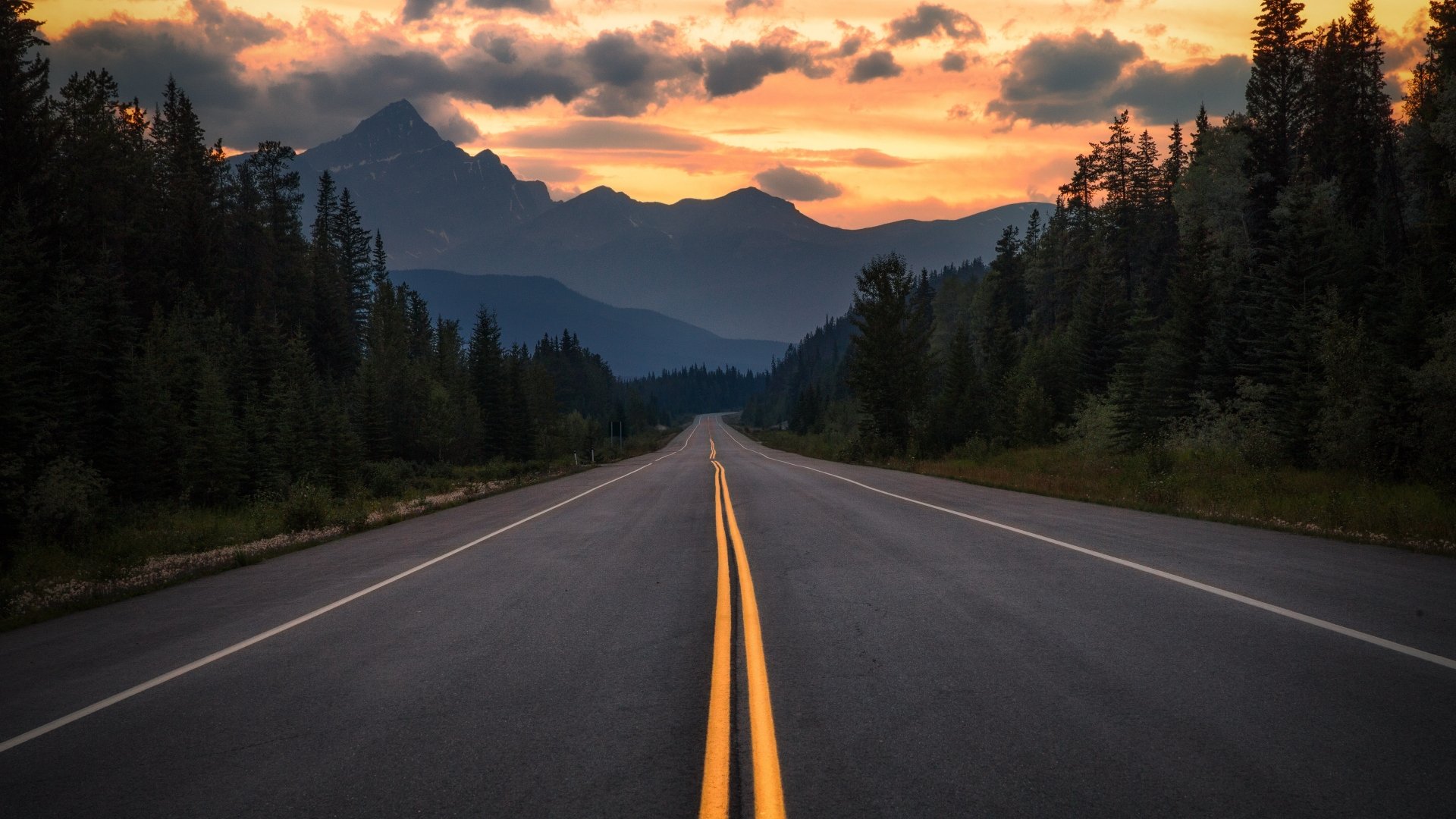 4K Ultra HD PC desktop wallpaper: man-made two-lane highway leading through pine forests toward silhouetted mountains under a fiery sunset.