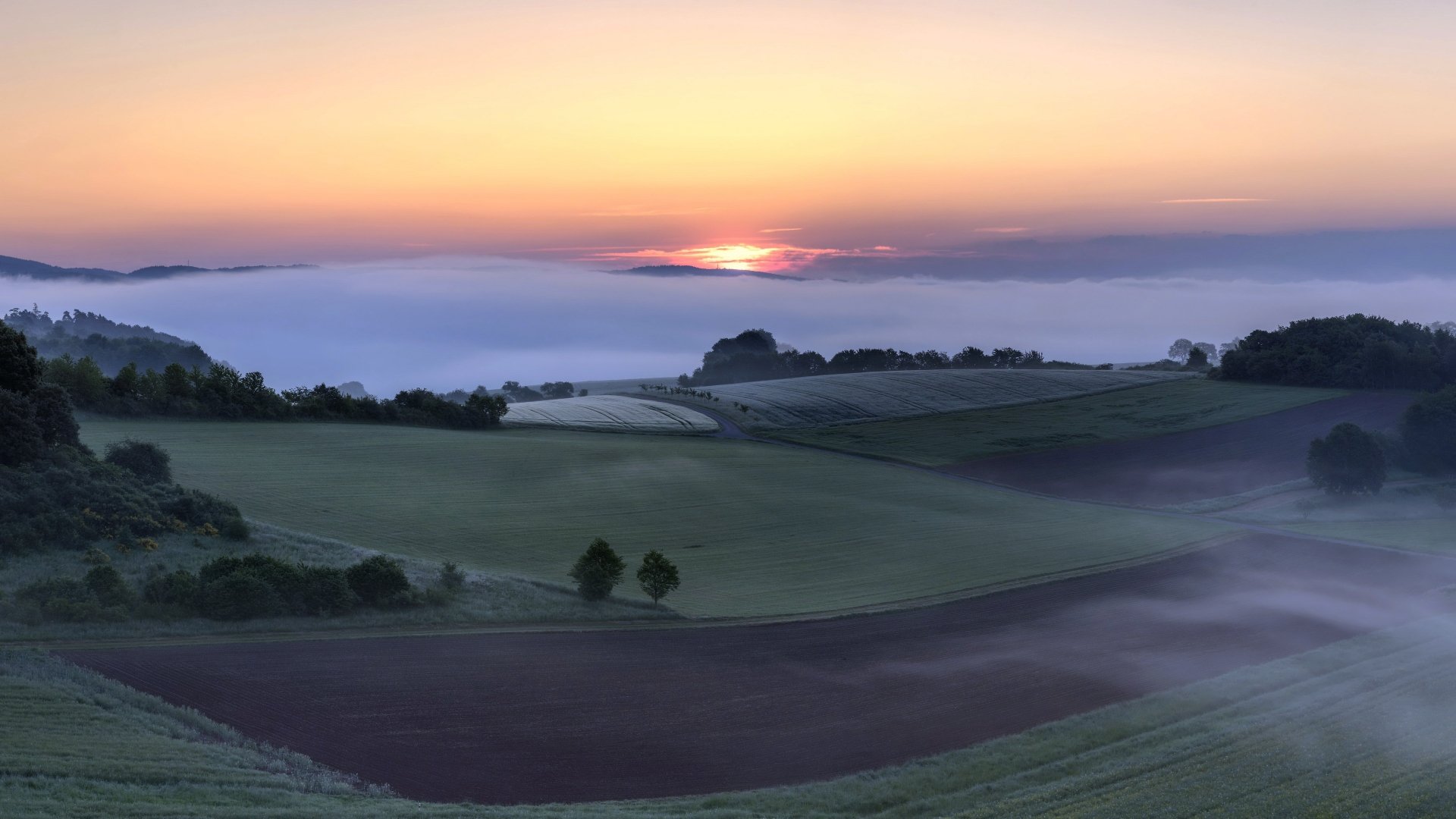 4K Ultra HD PC desktop wallpaper: misty rolling fields and patchwork farmland at sunrise, soft pastel sky above low-lying fog and gentle hills.