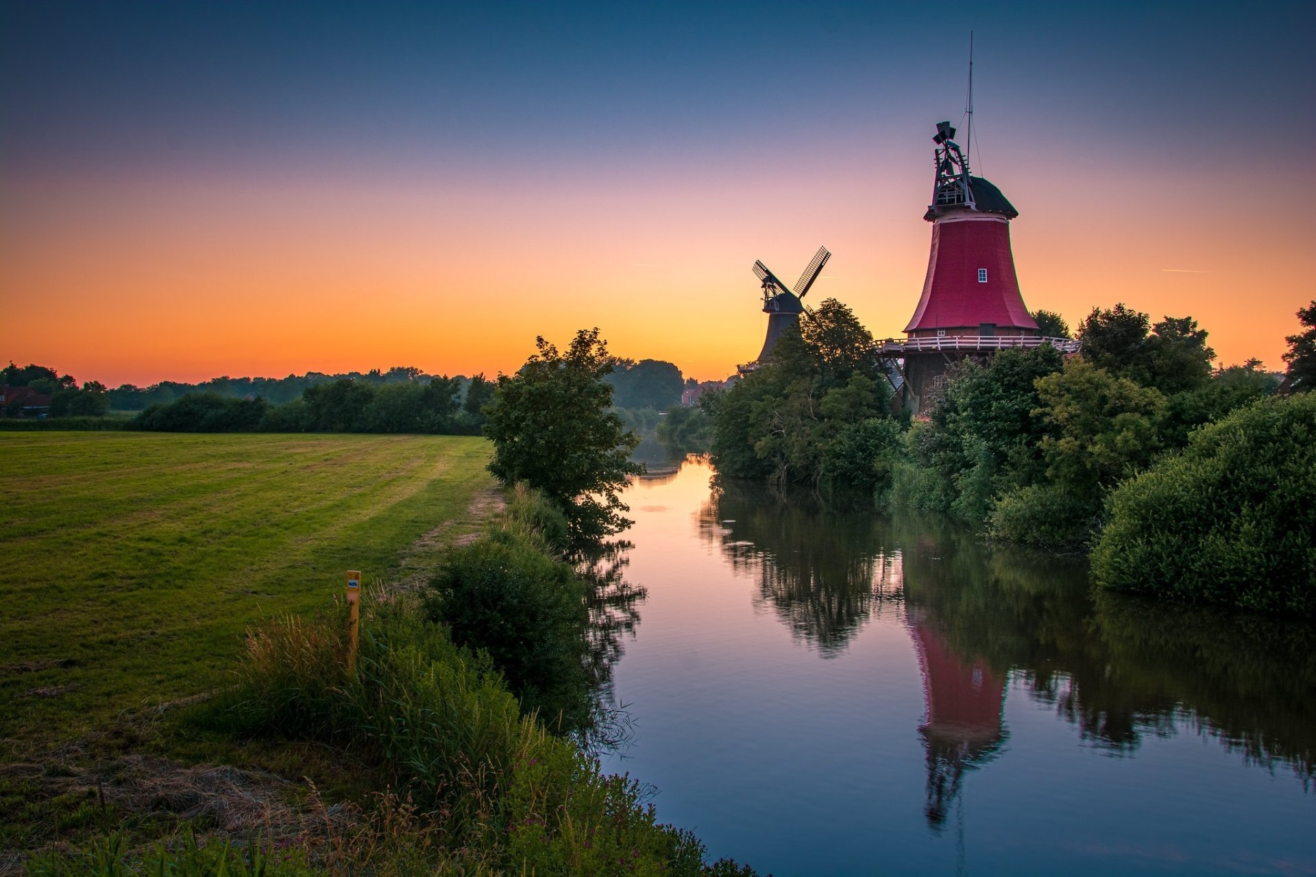 HD desktop wallpaper featuring a serene landscape with two man-made windmills reflected in a calm river at sunset.