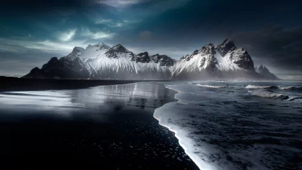 Vestrahorn Mountain rises dramatically above a dark beach in Iceland, with waves gently lapping the shore under a moody, cloud-filled sky in this 4K Ultra HD desktop wallpaper.