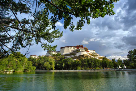 Scenic view of Tibet’s Potala Palace atop a mountain, overlooking a serene lake with lush greenery under a partly cloudy sky.