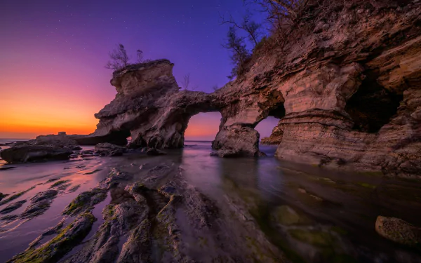 HD PC desktop wallpaper — nature coastal sunset with a stone arch, tide pools and reflective wet rock under a purple-orange sky.