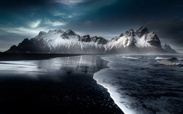 Vestrahorn Mountain rises dramatically above a dark beach in Iceland, with waves gently lapping the shore under a moody, cloud-filled sky in this 4K Ultra HD desktop wallpaper.