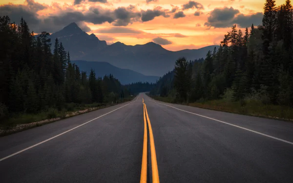 4K Ultra HD PC desktop wallpaper: man-made two-lane highway leading through pine forests toward silhouetted mountains under a fiery sunset.