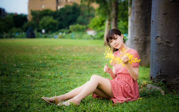 A brunette Asian woman in a pink dress sits on green grass against a tree, holding yellow flowers with a blurred natural background in 4K Ultra HD detail.