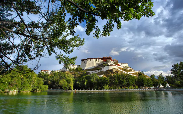 Scenic view of Tibet’s Potala Palace atop a mountain, overlooking a serene lake with lush greenery under a partly cloudy sky.