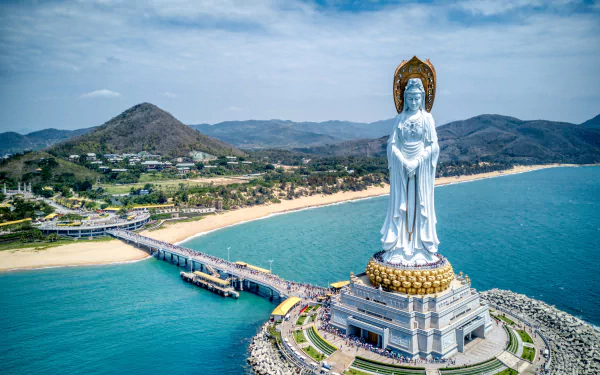 4K Ultra HD desktop wallpaper: aerial view of Nanshan Temple's towering Buddhist statue on a man-made coastal platform, arched bridge to shore, mountains, beach and landscape in China.