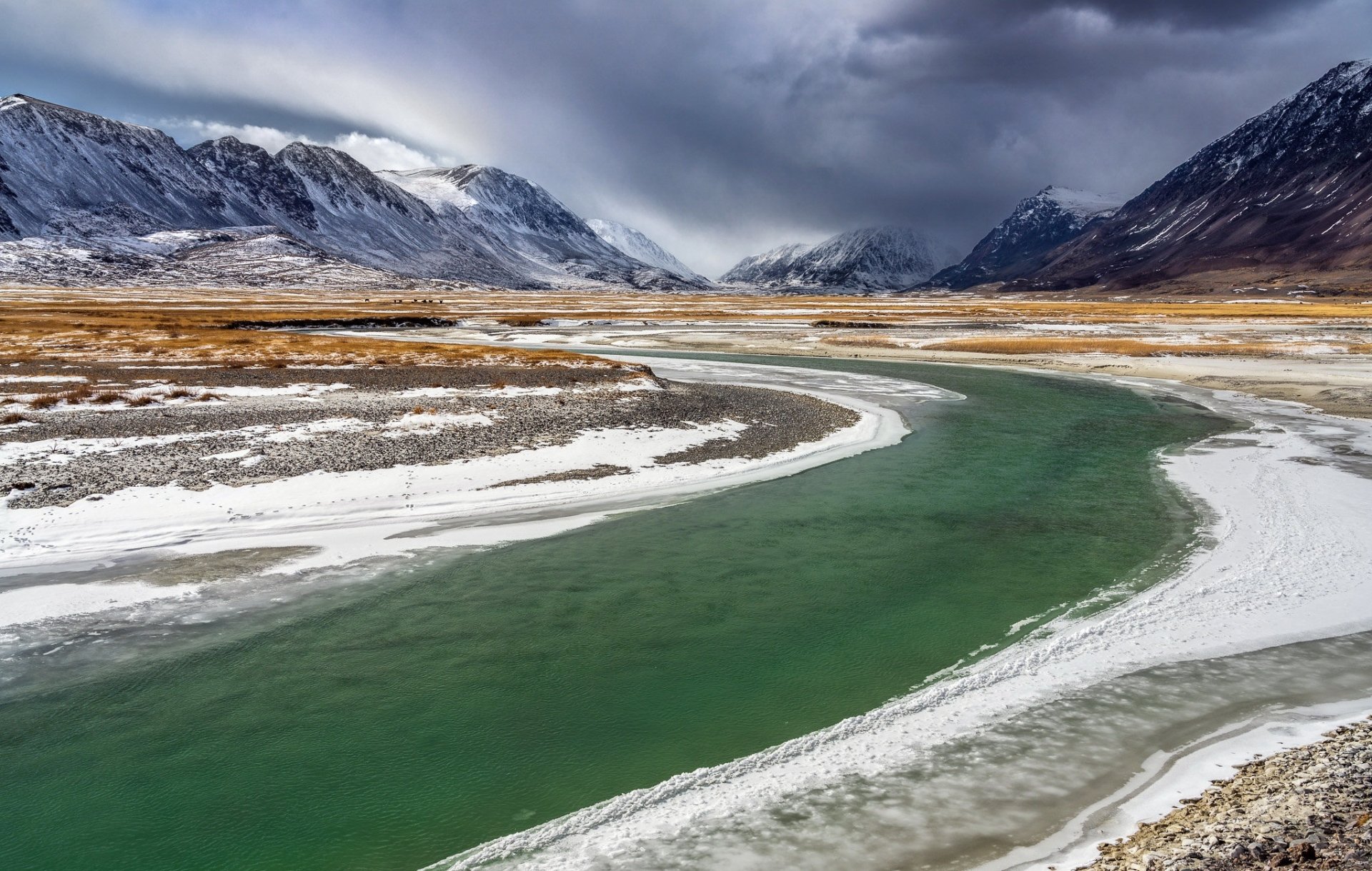 A winding green river flows through a snowy valley surrounded by towering mountains in Mongolia, showcasing the serene beauty of nature in HD quality.