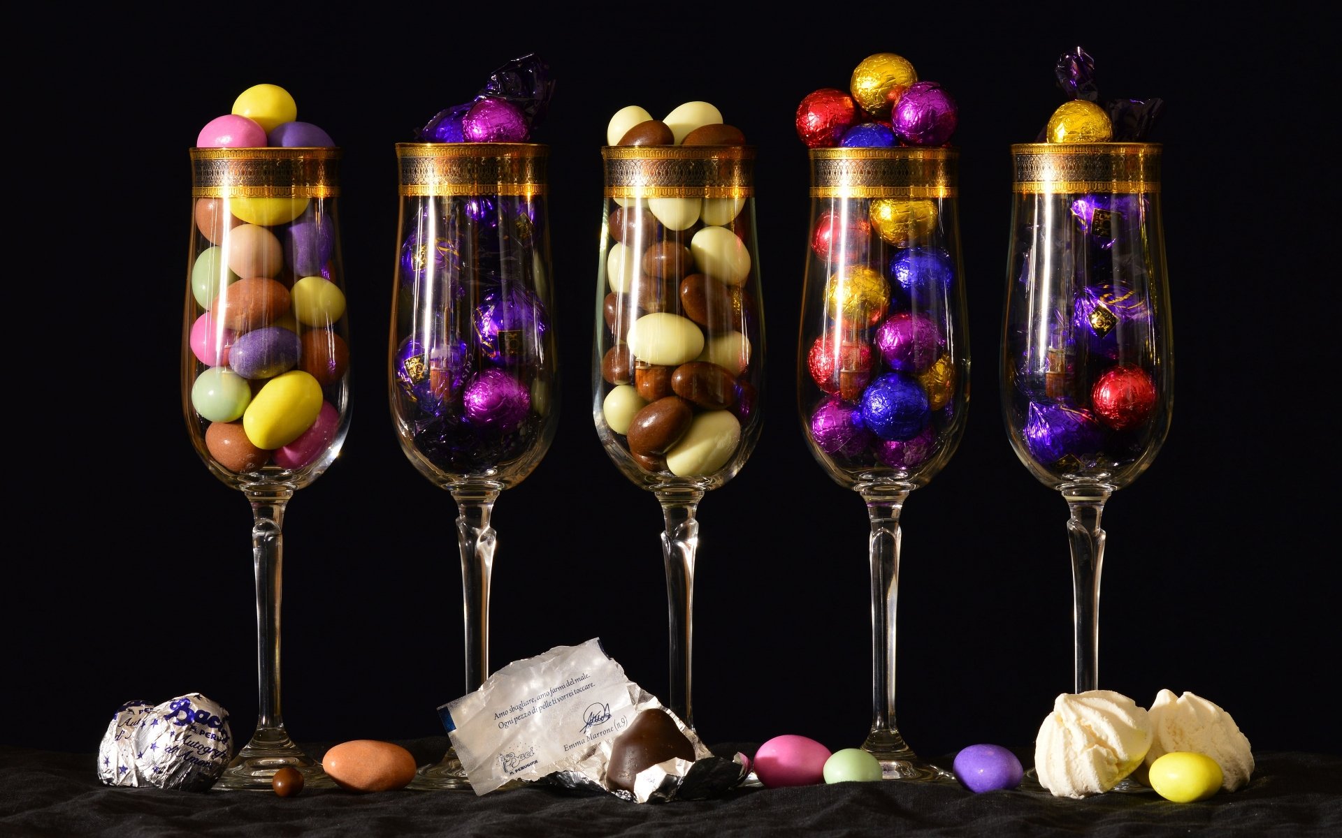 4K Ultra HD PC desktop wallpaper: five champagne flutes filled with colorful candies, foil-wrapped chocolates and meringues — confectionery food and scattered sweets on a black background.