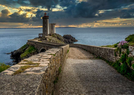 A 4K Ultra HD image of a man-made lighthouse on the rugged coast of Brittany, France, overlooking the sea under a dramatic cloudy sky at sunset.