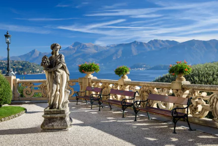 Villa Balbianello terrace at Lake Como, Italy: ornate balustrade, man-made classical statue and lined benches overlooking the lake and mountains — 2K Quad HD PC desktop wallpaper/background.