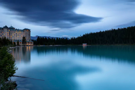 A serene view of Lake Louise in Alberta, Canada, featuring a grand hotel beside a calm lake surrounded by dense forest under a moody, cloudy sky.