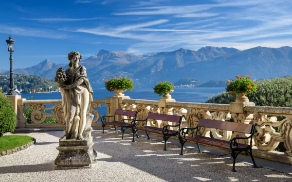 Villa Balbianello terrace at Lake Como, Italy: ornate balustrade, man-made classical statue and lined benches overlooking the lake and mountains — 2K Quad HD PC desktop wallpaper/background.