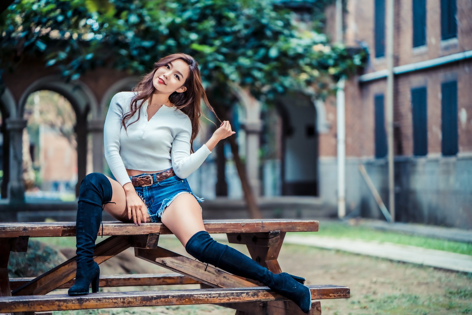 A brunette Asian woman model in shorts and knee-high boots sits on a wooden bench outdoors, captured with a shallow depth of field in 4K Ultra HD quality.