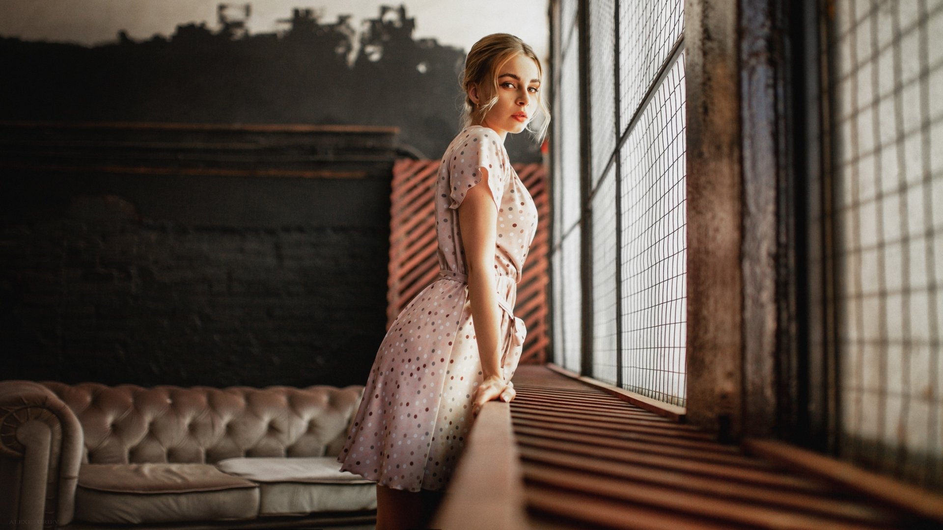 Blonde woman model in a light floral dress leans on a wooden railing by a large window in an industrial-style room, captured in a high-definition desktop wallpaper image.